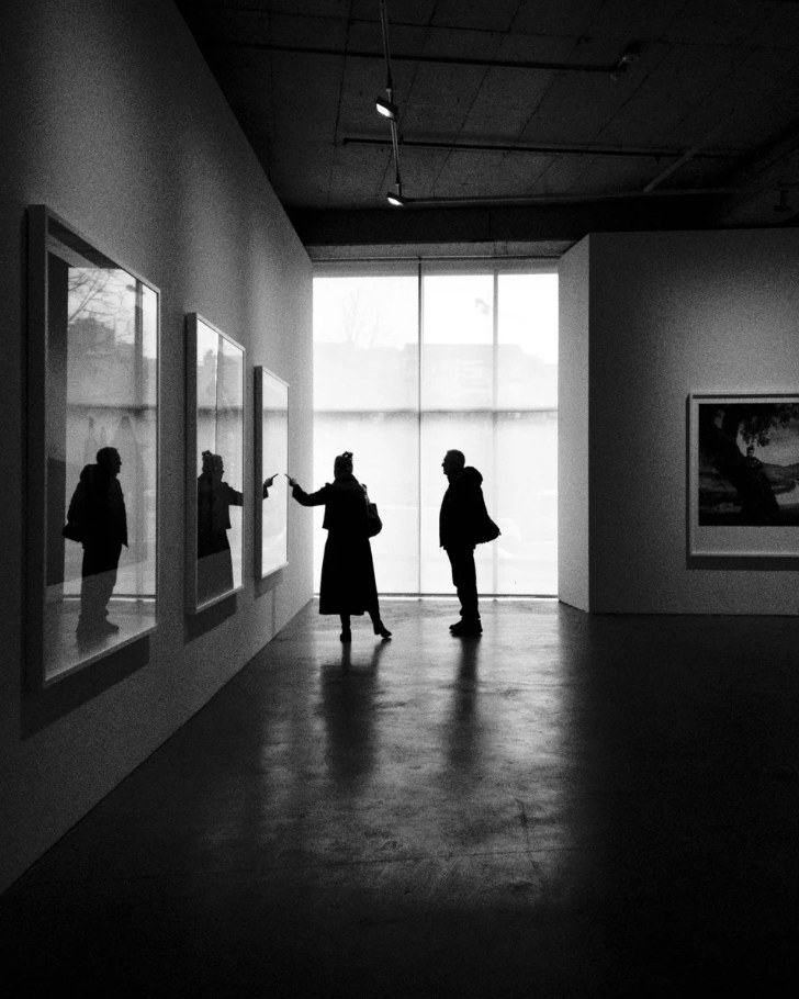Two silhouetted visitors stand in a dark gallery in front of a bright window, and one points at a framed photograph.